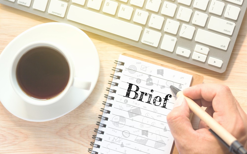 Image of a desktop with a computer keypad and a hand holding a pen to paper pad ready to write a brief